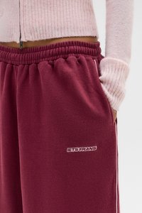 Bordo trenirke od mekanog materijala s elastičnim pojasom, bočnim džepovima i kontrastnim bijelim bočnim trakama. Sadrži izvezenu oznaku.
