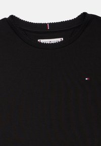 Czarny bawełniany t-shirt z okrągłym dekoltem wykończonym falbanką oraz małym logo w kolorach czerwonym, białym i niebieskim po lewej stronie.