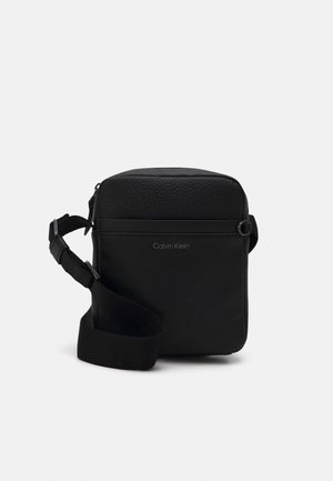 Czarna torebka na ramię z teksturowanej skóry, z regulowanym paskiem, zamkiem błyskawicznym oraz subtelnym logo "Calvin Klein" na przodzie.