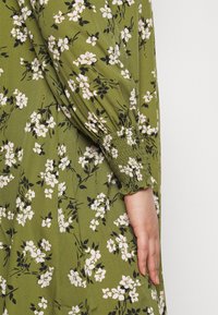 Abito floreale verde con motivi di fiori bianchi e neri, con maniche smock e morbido tessuto, mostrato di lato.