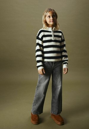 Gestreifter Pullover in Schwarz und Weiß mit einem halben Reißverschluss-Kragen, kombiniert mit weit geschnittenen grauen Jeans und braunen Wildleder-Plateau-Stiefeln.