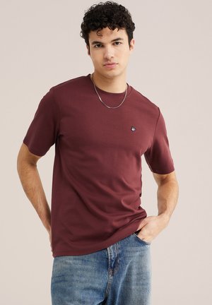 Fiatal férfi göndör hajjal, bordó pólót és kék farmert visel, egyik kezével a zsebében áll, egyszínű háttér előtt.