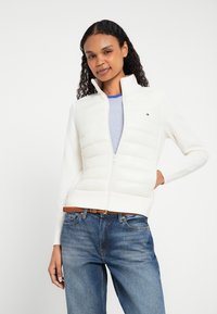 Λευκό μπουφάν με φερμουάρ, με καπιτονέ μπροστινή όψη, υφασμένα μανίκια και ψηλό γιακά. Φορεμένο πάνω από μπλε ριγέ πουκάμισο και συνδυασμένο με μπλε τζιν.