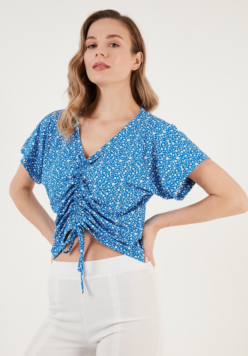 Top avvolgente floreale blu con maniche corte e dettaglio con stringa in vita, realizzato in tessuto leggero. Il motivo include piccoli accenti bianchi.