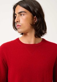 Maglione rosso lavorato a maglia con scollo a girocollo e polsini a costine. Presenta una texture liscia e un accento triangolare a costine sulla cucitura della spalla.