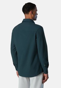 Camisa de manga larga en verde oscuro con cuello, que presenta puños abotonados y una textura suave. Líneas limpias con detalles de diseño mínimos.