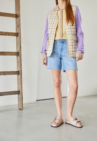 Beige kviltad väst över en gul topp, ljusblå denimshorts och beige slidesandaler med en dekorativ spänne. Texturerade material, avslappnad stil.