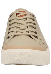 Beige canvas sneakers with flat laces, rounded toe, rubber sole, and textured detailing. Brown leather accent at the tongue.
