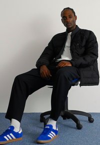 Schwarze Puffjacke über einem weißen Hemd, kombiniert mit schwarzen Hosen und blauen Adidas-Sneakers. Der Hintergrund ist rein weiß mit einem blauen Teppich.