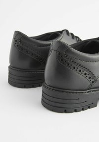 Pantofi negri din piele, cu un design brogue detaliat, formă rotundă, suprafață texturată și o talpă groasă din cauciuc cu striații.