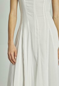 Weißes Kleid mit vertikalen Falten und strukturiertem Oberteil. Texturiertes Gewebe, ausgestellter Rock und taillierte Passform. Detail einer Hand, die auf dem Rock ruht.