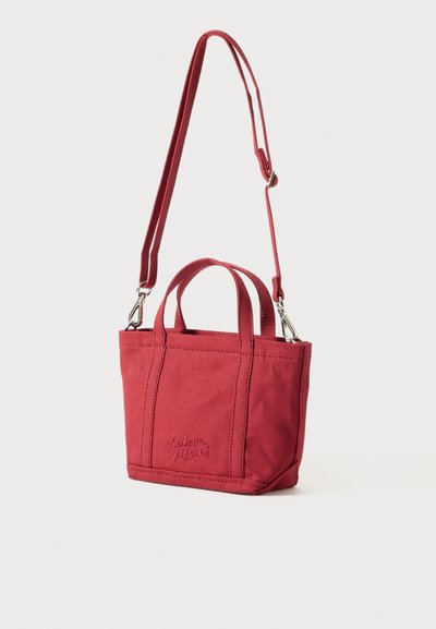 Saco de tela vermelho com duas alças curtas e uma alça destacável. Apresenta detalhes de costura e um logótipo em relevo na parte da frente.