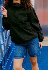 Personne aux cheveux bouclés portant une veste noire et un short en jean bleu s’appuie contre une clôture en métal bleue sur un sol bleu.
