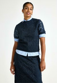 Marineblå blonde top med korte ærmer over en lyseblå stribet skjorte med krave. Den har et detaljeret blomsterdesign og en tætsiddende pasform med rund halsudskæring.