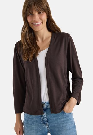 Femme souriante portant un cardigan marron foncé ouvert sur un top blanc et un jean bleu, la main droite dans sa poche.