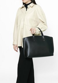 Zwarte leren tote bag met een gladde textuur en een afgeronde bovenkant, voorzien van dubbele handvatten en messing drukknoopaccenten, vastgehouden door een persoon in neutrale kleding.