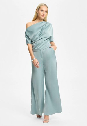 Donna bionda che indossa un top azzurro chiaro di seta senza spalle e pantaloni a gamba larga, accessoriata con braccialetti e tacchi aperti, in piedi.