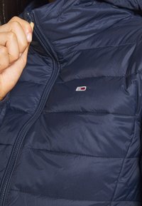 Marinblå pufferjacka med en glänsande finish, horisontellt quiltat mönster och en dragkedjestängning. Har en liten, märkeslogotyp på framsidan.