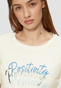 Crème-kleurig T-shirt met een ronde halslijn, voorzien van blauwe en zilveren grafische tekst "Positivity" in verschillende stijlen en gewichten.