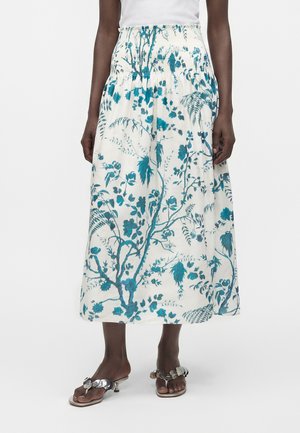Vue de la taille aux chevilles d'une personne portant une jupe midi blanche à imprimé floral bleu et des sandales à talons argentées avec des décorations circulaires.