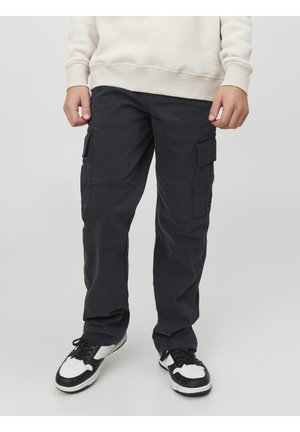 Personne portant un pantalon cargo noir avec poches latérales et des baskets noir et blanc, debout devant un fond uni.
