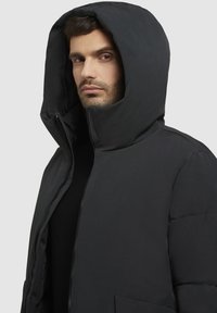 Crna puffer jakna s velikim kapuljačom, s prednim zatvaračem i bočnim džepovima. Tkanina je glatka s laganim sjajnim završetkom.