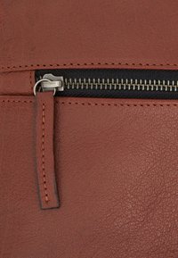 Smeđa kožna torba s zip-zipper džepom. Ima teksturiranu površinu, metalni zip i tkaninsku podstavu koja se vidi uz rub zipa.