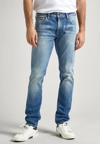Vaqueros de mezclilla de color azul claro con un acabado desgastado, corte recto, cinco bolsillos y una etiqueta de marca visible en la cinturilla. Zapatillas blancas complementan el look.