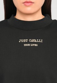 Felpa nera con colletto alto, con la scritta "JUST CAVALLI TRUE LOVER" ricamata in beige. Il testo è centrato e ben visibile.