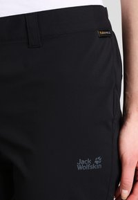 Černé elastické kalhoty od Jack Wolfskin, se hladkým povrchem, štítkem Flexshield a nenápadným logem poblíž pasu.