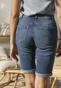 Personne portant un short en jean roulé, debout à l'intérieur près de meubles en osier avec des coussins dans une lumière naturelle douce.
