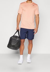 Pánský sportovní outfit s krátkým rukávem v jemně růžové barvě, tmavě modré šortky a černá sportovní taška. Bílí tenisky doplňují celý vzhled.