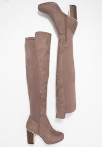 Botas marrones por encima de la rodilla con material de ante, detalle de cremallera lateral y tacón grueso. Acabado suave con diseño de puntera redondeada.