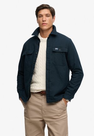 Mand iført en marineblå jakke over en hvid kabelstrikket sweater og beige bukser med hænderne i lommerne, stående mod en ensfarvet baggrund.