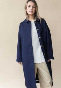 Marineblauer Mantel mit großen Knöpfen, spitzem Kragen und seitlichen Taschen; getragen über einem weißen Strickpullover und beigen Hosen. Glatte Stofftextur.
