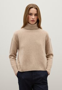 Beige Wollrollkragenpullover mit einem leicht übergroßen Schnitt, geripptem Kragen und Bündchen, kombiniert mit dunkelblauen Hosen. Glatte Textur.