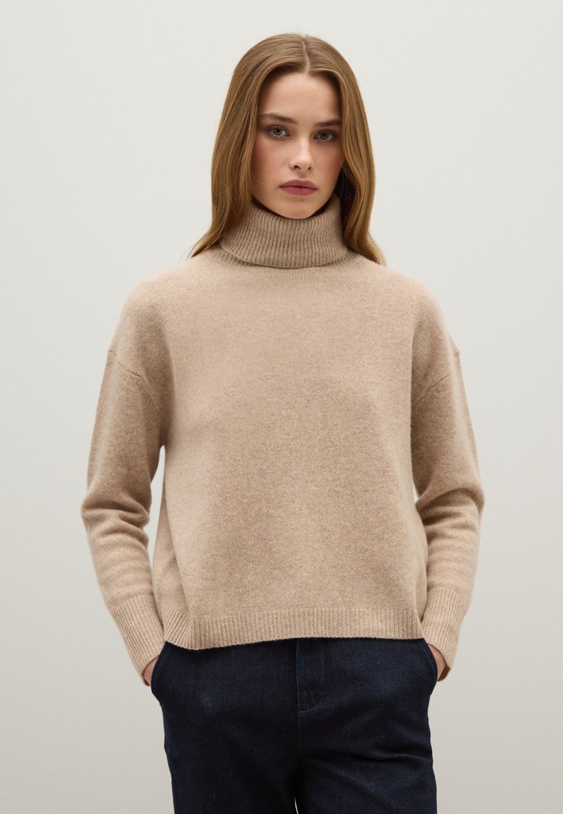 Beige Wollrollkragenpullover mit einem leicht übergroßen Schnitt, geripptem Kragen und Bündchen, kombiniert mit dunkelblauen Hosen. Glatte Textur.