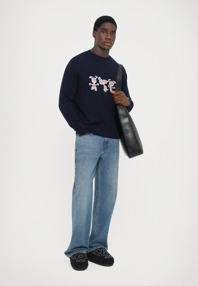 Pulover navy cu un model decorativ în roz și negru, asortat cu blugi albaștri deshi și pantofi negri slip-on, ținând o geantă neagră.