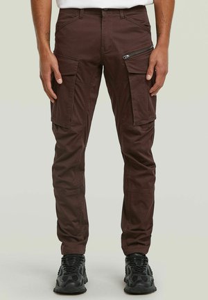Homme portant un pantalon cargo marron foncé avec plusieurs poches et des baskets noires, debout devant un fond clair uni.
