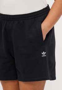 Juodi šortai, pagaminti iš medvilnės ir sintetinių pluoštų mišinio, su elastiniu juosmeniu, šoninėmis kišenėmis ir baltu "Adidas" logotipu kairėje apatinėje pusėje.