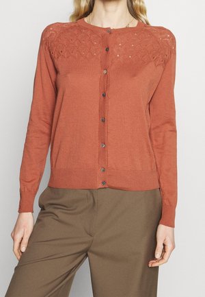 Femme portant un cardigan boutonné couleur rouille avec un détail en dentelle sur les épaules et un pantalon marron sur fond uni