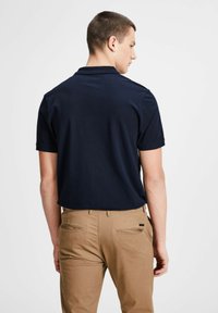 Navyblaues Poloshirt mit kurzen Ärmeln, glatter Textur, klassischem Kragen, getragen mit beigen Hosen mit Taschen auf der Rückseite und subtiler Detailverarbeitung.