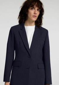 Mørkeblå skreddersydd blazer med hakkeslag, to knapper og sidelommer. Laget av strukturert stoff, har en enkelt brystlomme.