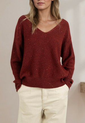 Pull-over en tricot rouge à col en V avec une finition texturée et de subtils accents pailletés, associé à un pantalon beige clair. Coupe ample et poignets côtelés.