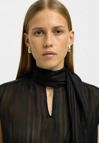 Blouse noire transparente à rayures verticales avec un décolleté en forme de trou de serrure, assortie d'une écharpe en tissu assorti. Le mannequin porte des boucles d'oreilles créoles argentées.