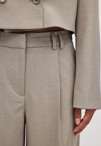 Costume taillé gris clair avec une veste courte, ornée de grands boutons et de pantalons fuselés. Tissu lisse avec une texture subtile et une coupe structurée.