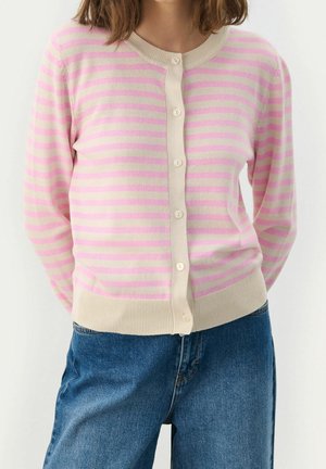 Femme portant un cardigan rayé rose et crème boutonné et un jean denim bleu, debout les mains derrière le dos.
