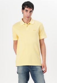 Gul polo skjorte laget av bomull, med krage, kortermet, og et lite brodert logo på brystet. Brukt sammen med blå jeans.