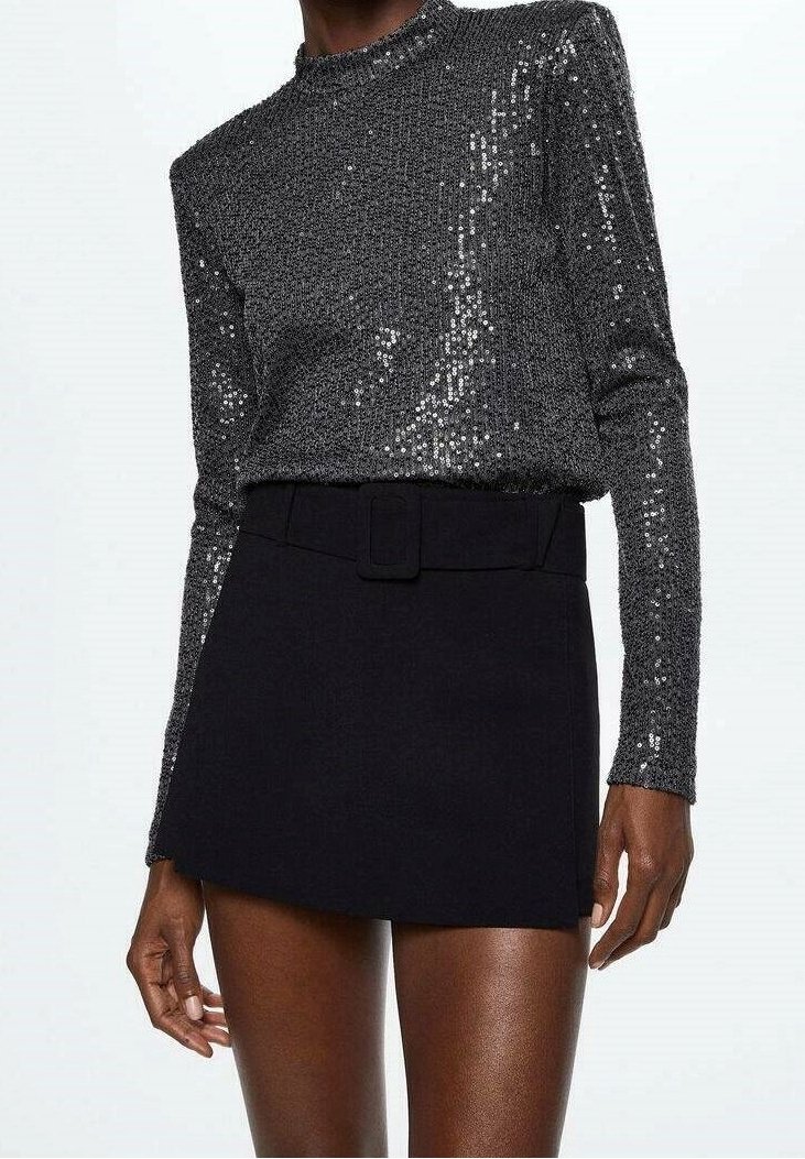 Femme portant un haut à paillettes gris foncé à manches longues rentré dans une mini-jupe noire taille haute avec une large ceinture.