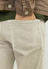 Jeans beige con una tela texturizada, que presentan un bolsillo trasero con detalles de costura sutiles. Combinados con una camisa verde oscura y una chaqueta marrón.